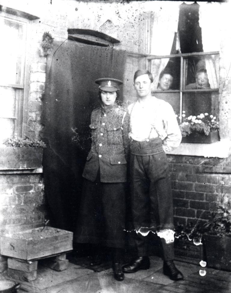 john with his girlfriend. she is wearing his cap and jacket and they are standing outside beside the back door  of a house. two people are looking through a window at them