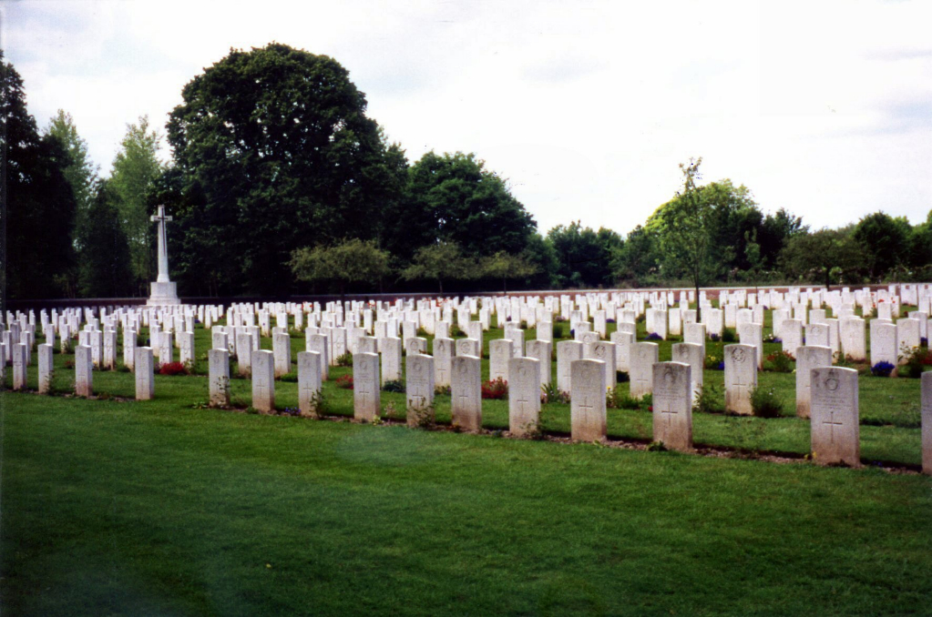 Rows of white gravestones dominate the photograph with a stone memorial cross in the background