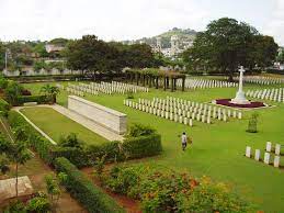 an aerial photograph of the cemetery with the white headstones laid out in rows surrounding the white cross of sacrifice on the right, on the left is a white stone plinth with a hedge and trees