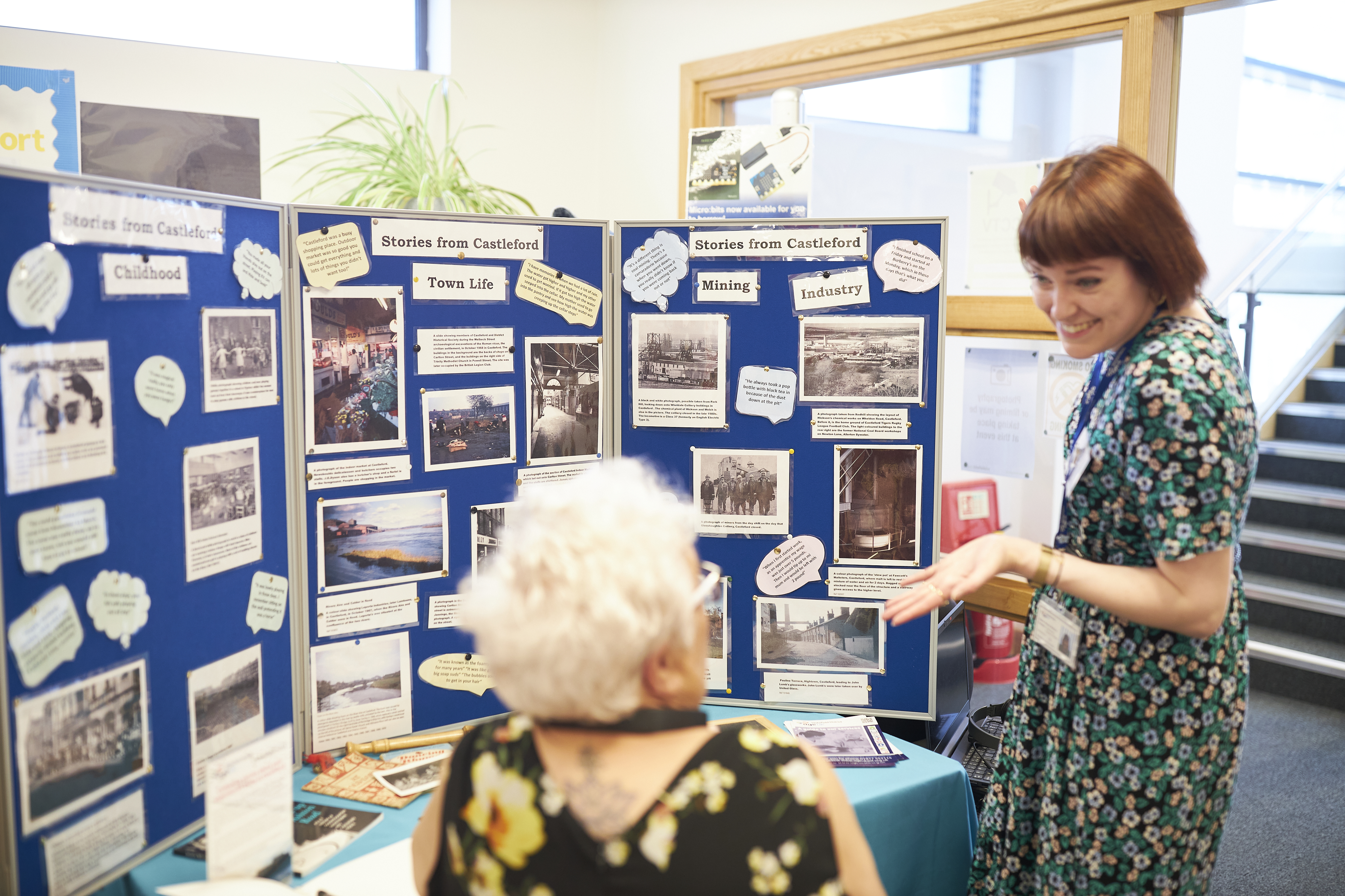 A standing person is talking to a seated person in front of a community heritage display